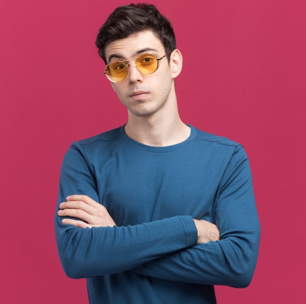 confident young brunette caucasian boy in sun glasses standing with crossed arms isolated on pink background with copy space